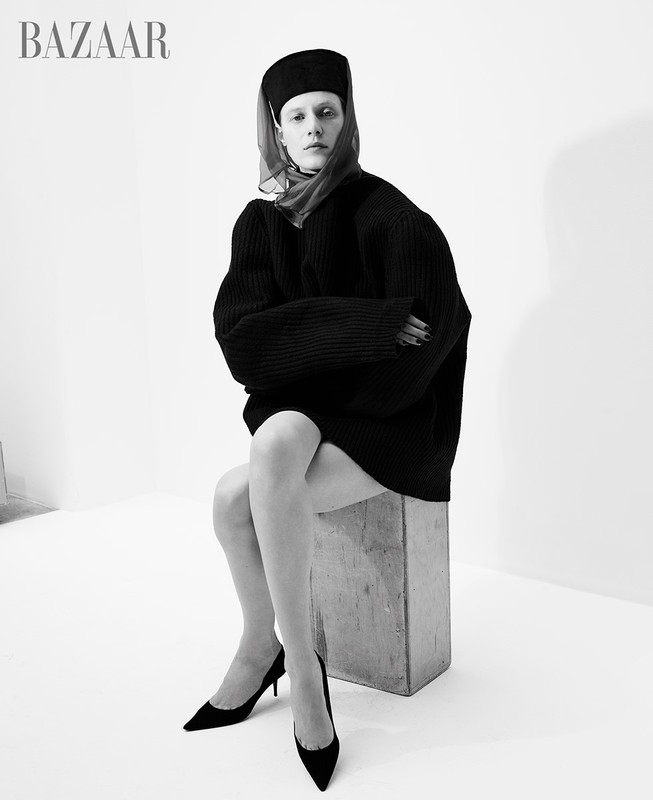 Monochrome fashion shot of a woman with a dark headscarf and oversized sweater, seated on a concrete block, wearing high heels in a white studio.