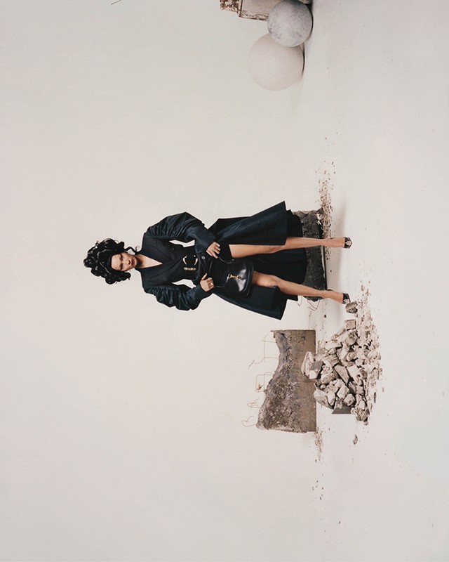 A woman in a black coat and heels poses amid a white background with concrete rubble. Fashion photography, stylish outerwear.