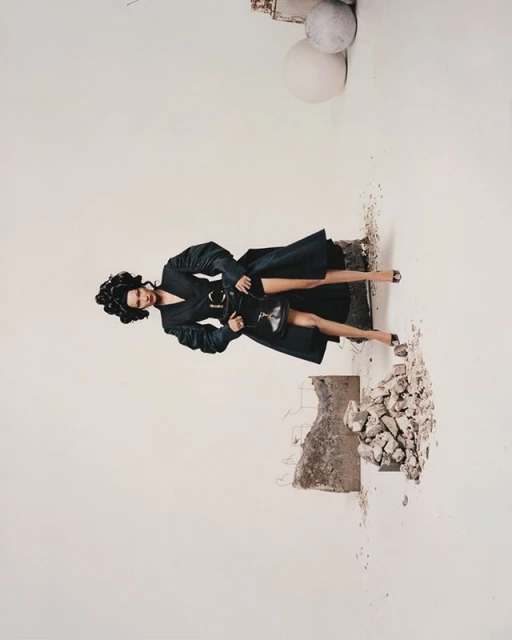 A woman in a black coat and heels poses amid a white background with concrete rubble. Fashion photography, stylish outerwear.