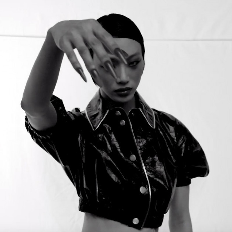 Striking black and white portrait of a stylish woman with long nails and a shiny jacket, posed against a plain white backdrop.