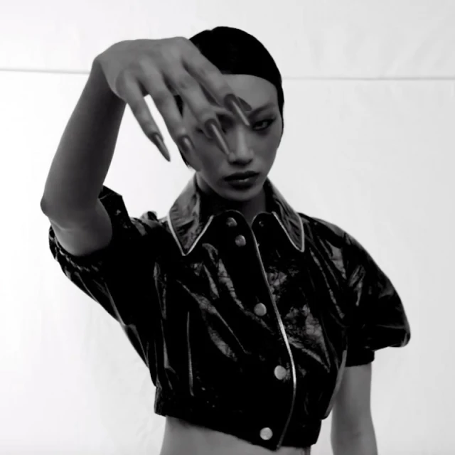 Striking black and white portrait of a stylish woman with long nails and a shiny jacket, posed against a plain white backdrop.