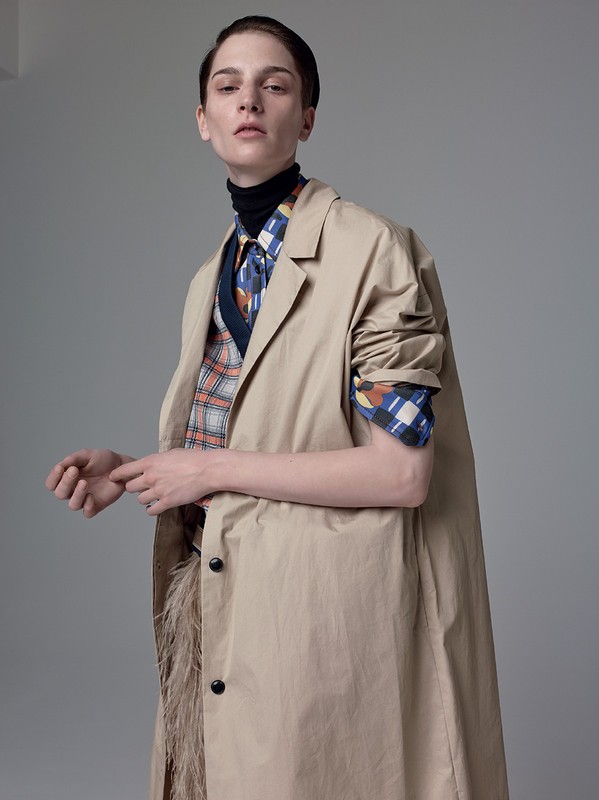 A woman in a beige trench coat over a black turtleneck, patterned shirt and vest poses in a fashion studio portrait with neutral background.