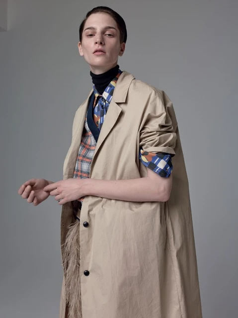 A woman in a beige trench coat over a black turtleneck, patterned shirt and vest poses in a fashion studio portrait with neutral background.