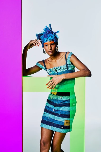 Fashion model wearing a blue denim-style dress with lime and magenta accents, posing in a studio with a blue hairstyle.