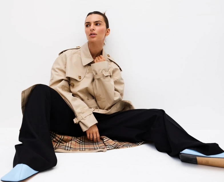 Adult female model wearing a beige trench coat and black pants sits in a bright, white photo studio looking away from the camera.