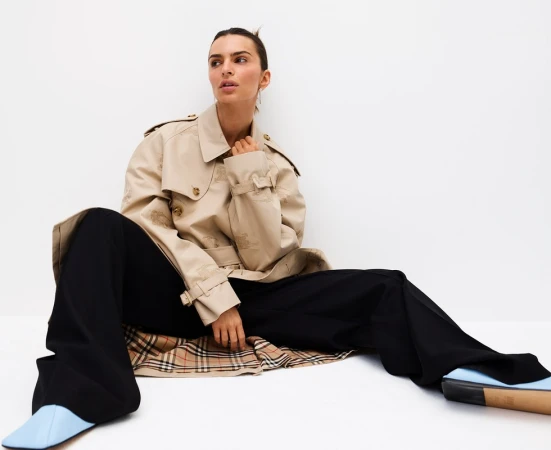 Adult female model wearing a beige trench coat and black pants sits in a bright, white photo studio looking away from the camera.