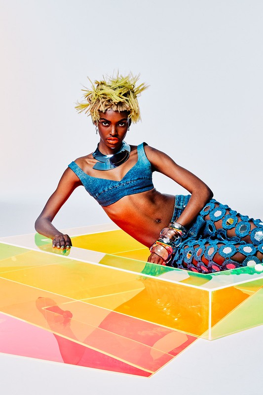 Young Black woman with spiky blonde hair poses in a denim crop top and cut-out pants on a colorful geometric block in a white studio.
