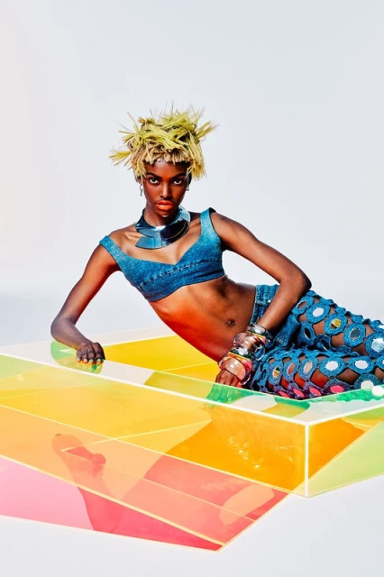 Young Black woman with spiky blonde hair poses in a denim crop top and cut-out pants on a colorful geometric block in a white studio.
