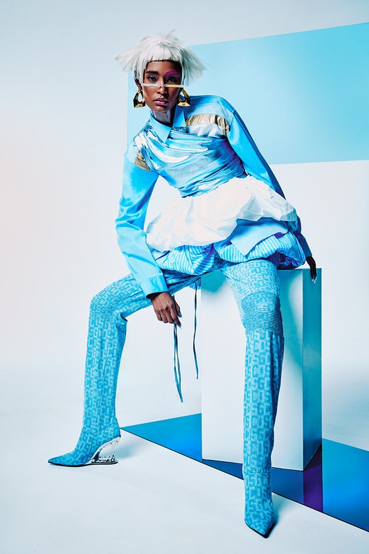 A person with a white wig and gold glasses poses seated on a white block wearing a light blue satin shirt and patterned leggings.