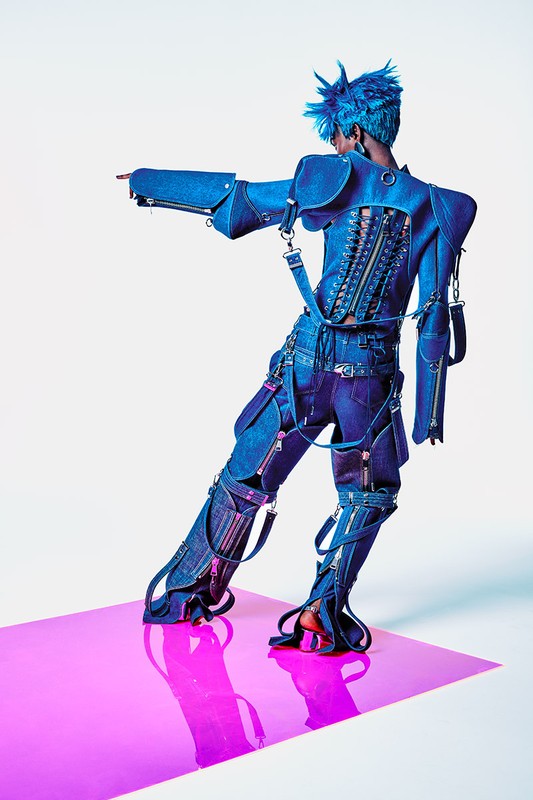 A person wearing a blue denim outfit with spiky blue hair stands on a pink surface in a studio, pointing their left hand to the side.