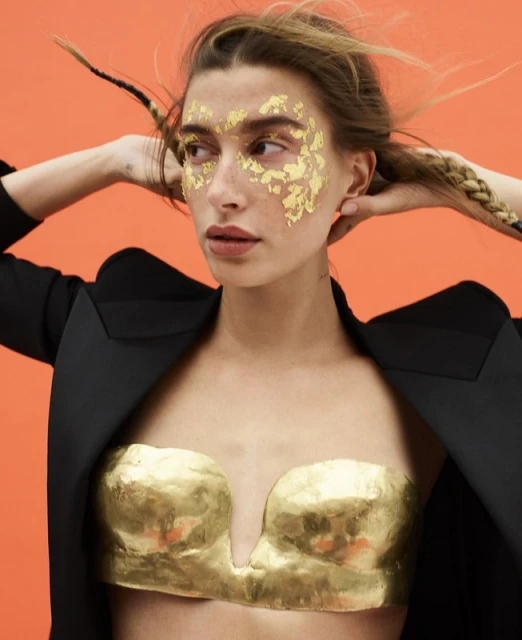 Portrait of an adult woman with gold foil on her face wearing a black blazer and a gold bra top in front of an orange background.