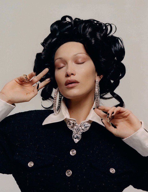Close-up portrait of model Bella Hadid with dark voluminous curly hair, large crystal hoop earrings, and black fingernails against a neutral background.