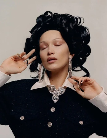Close-up portrait of model Bella Hadid with dark voluminous curly hair, large crystal hoop earrings, and black fingernails against a neutral background.