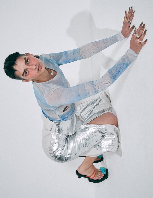 A person with short dark hair wearing a patterned top, metallic skirt, and sandals posing in front of a white background in a studio setting.
