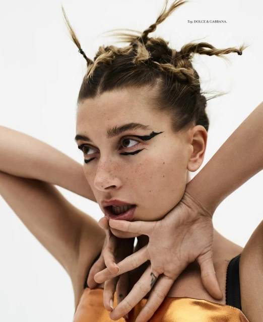 Close-up of a young woman with braided hair and heavy black winged eyeliner, wearing an orange sleeveless top against a white backdrop.