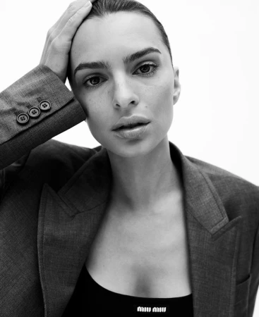 Close-up portrait of Emily Ratajkowski wearing a grey blazer and black top with hair pulled back, against a plain white background.