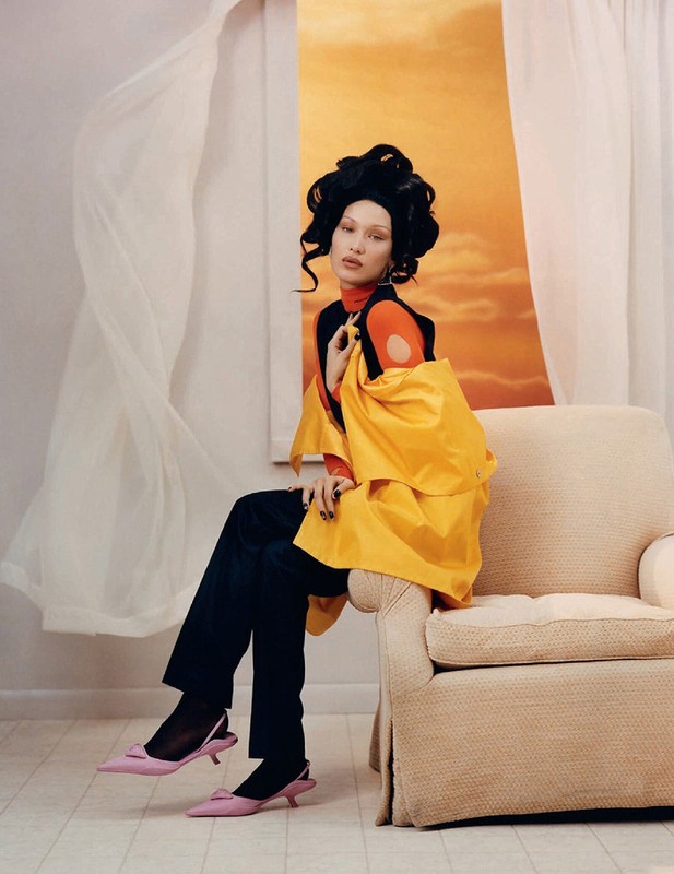 A woman with styled hair sits on a beige armchair wearing an orange turtleneck, black vest, yellow coat, black pants, and pink heels.