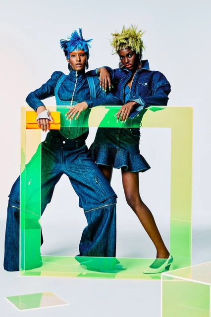 Two fashion models in denim outfits pose behind a translucent green and yellow rectangular frame in a studio, showcasing modern style.