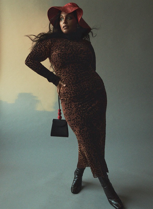 Portrait of a plus-size Black female model wearing a patterned dress, red hat, and ankle boots against a neutral studio backdrop.