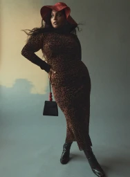 Model in animal print dress and red hat posing against grey wall with black handbag