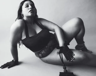 Plus-size model in a dark top and patterned bottoms posing on a seamless light background with black gloves.
