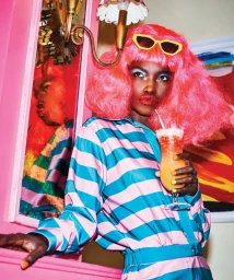 Model with vibrant pink wig and yellow sunglasses holding a cocktail in front of a pink wall and mirror