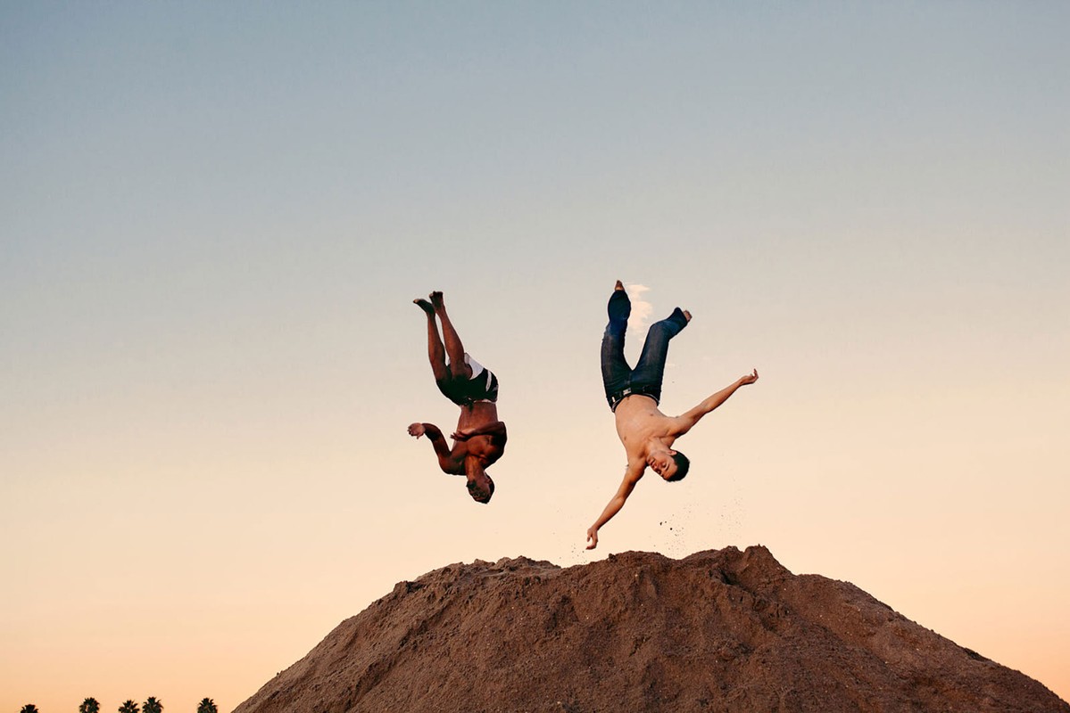 Two shirtless adult male athletes perform backflips on a dirt hill under a clear, gradient sky at golden hour; fitness and sport.