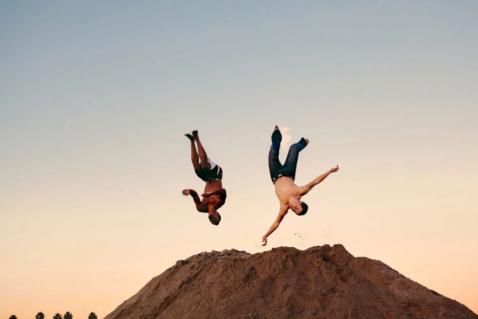 Two shirtless adult male athletes perform backflips on a dirt hill under a clear, gradient sky at golden hour; fitness and sport.