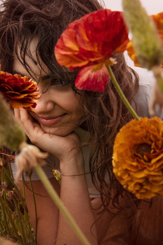 Close up of a smiling woman with curly hair resting her face on her hand surrounded by orange and red ranunculus flowers.