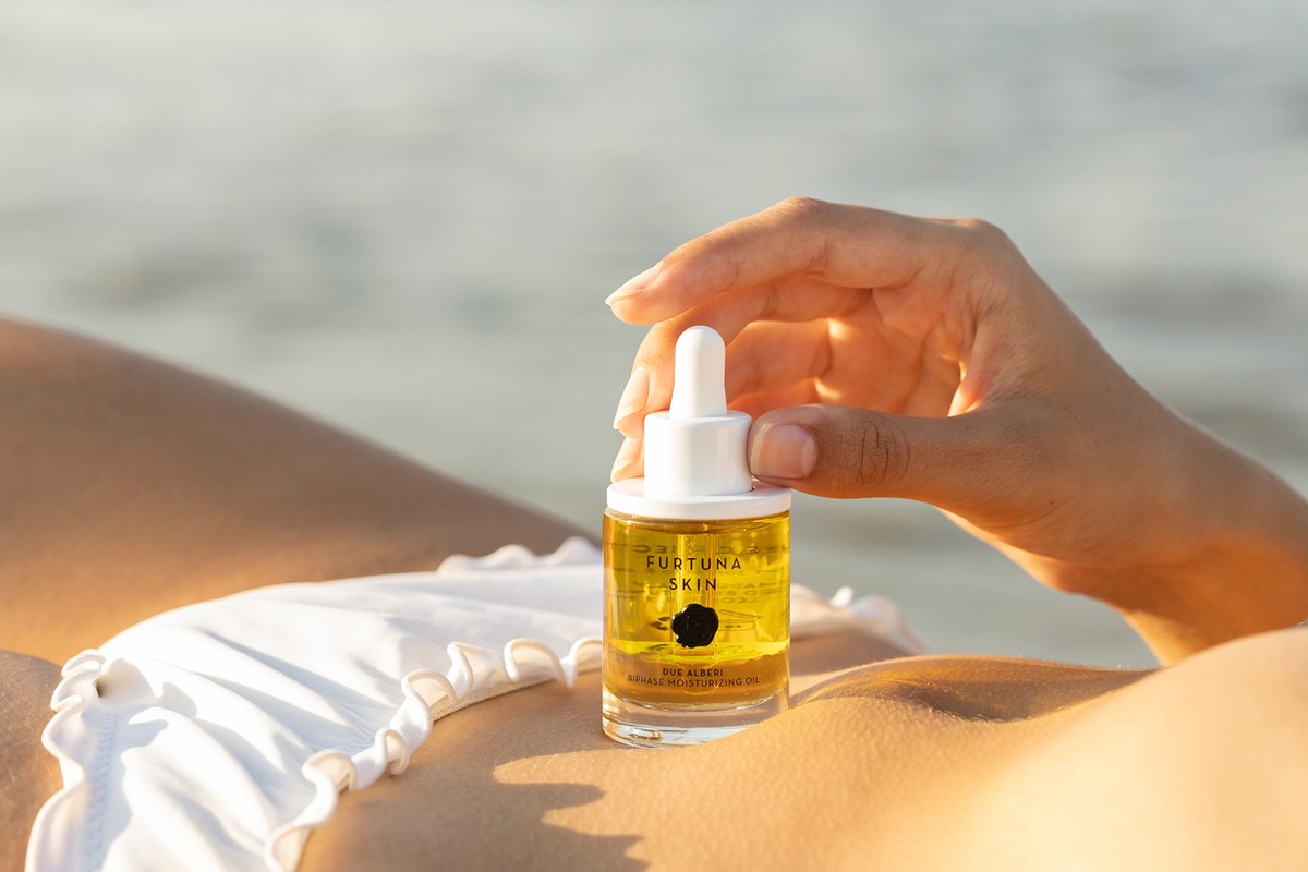 Close-up of a woman's hand holding a bottle of Eurtuna Skin face oil on the beach, showcasing skincare in a natural setting.