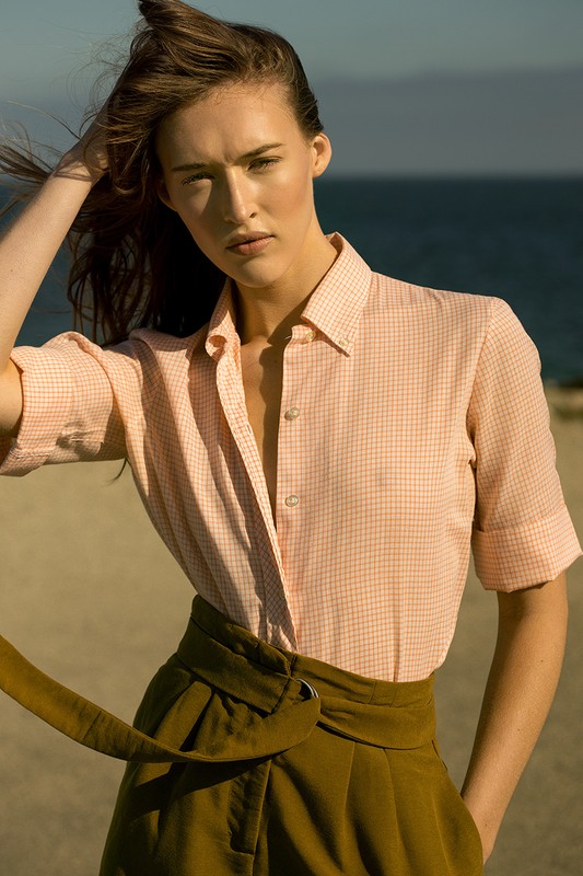 Model in peach-checkered button-down shirt and high-waisted olive trousers standing on a sandy beach.