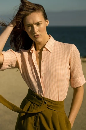Model in peach-checkered button-down shirt and high-waisted olive trousers standing on a sandy beach.