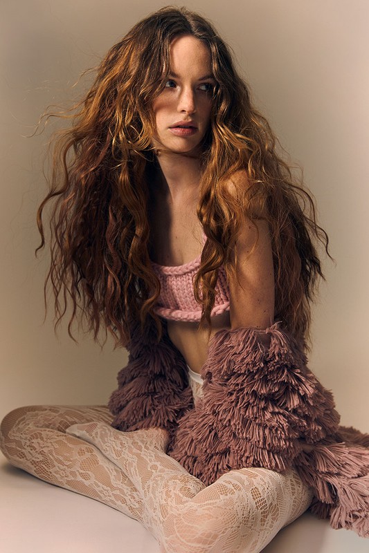 Woman with long curly hair wearing a pink crop top and furry shrug sits cross-legged in a studio with neutral background.