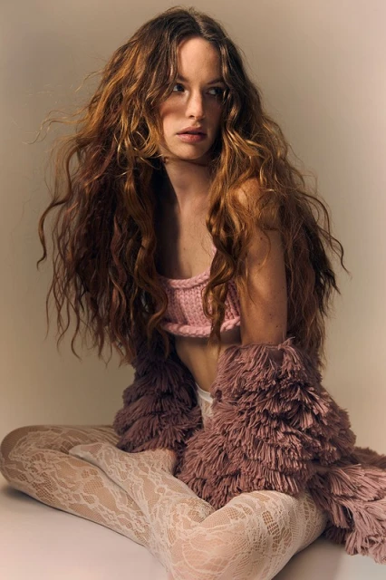 Woman with long curly hair wearing a pink crop top and furry shrug sits cross-legged in a studio with neutral background.