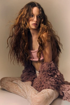 Woman with long curly hair wearing a pink crop top and furry shrug sits cross-legged in a studio with neutral background.