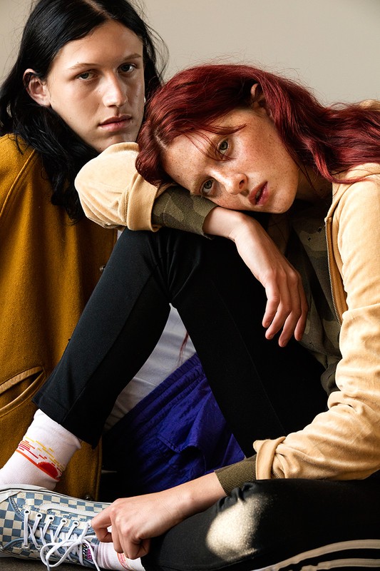 Two people sit indoors: A red-haired woman leans on a dark-haired person in a tan jacket. They wear casual clothing, sneakers, and colorful socks.