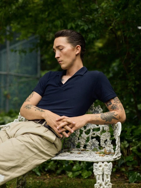 Man with tattoos poses on a white iron bench seat in a green backyard in a dark blue polo shirt and khaki pants with hair slicked back.