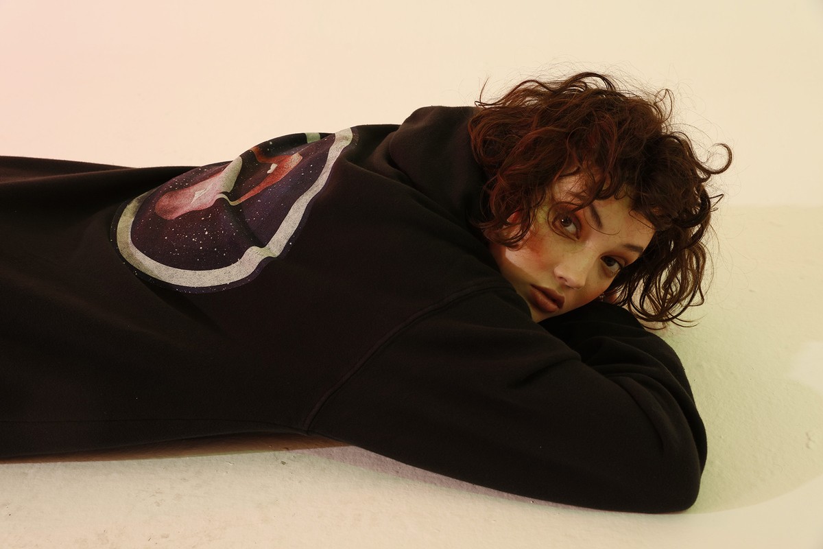 Adult female with auburn curly hair wearing a black hoodie with an art print, lying on her stomach in a brightly lit indoor setting.