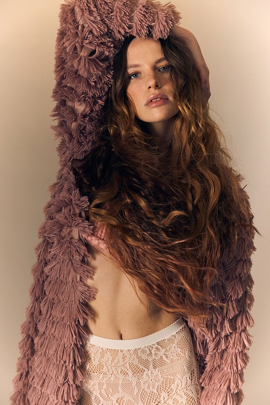 A woman in a fur coat and white lace shorts poses indoors in a medium studio shot with a neutral background.
