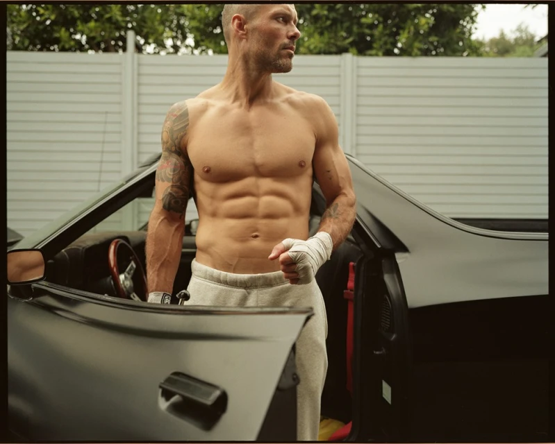 Shirtless man with tattoos and hand wraps stands by a black car outdoors, looking off to the side against a white fence backdrop.