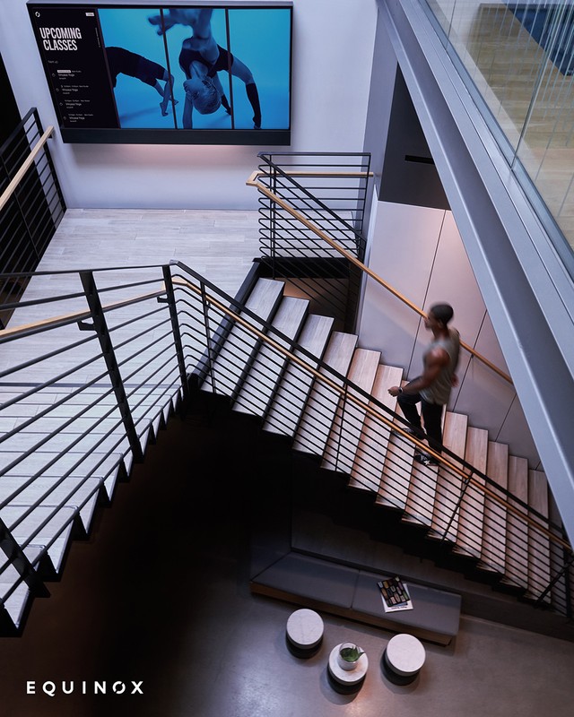 Interior view of Equinox fitness center with stairs, a person walking up, and a screen displaying exercise classes.