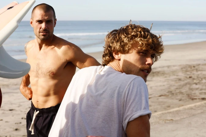 Two shirtless men standing on a sandy beach near an ocean with a surfboard in the foreground.