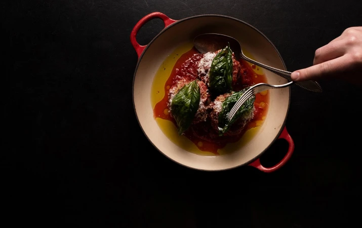 Overhead shot of meatballs in red sauce, garnished with basil, in a red-handled beige pan, alongside a fork and spoon, on a black surface.