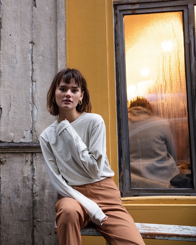 Young adult female with bangs and long-sleeved shirt posing outdoors against concrete wall and yellow-framed window reflecting sunset.