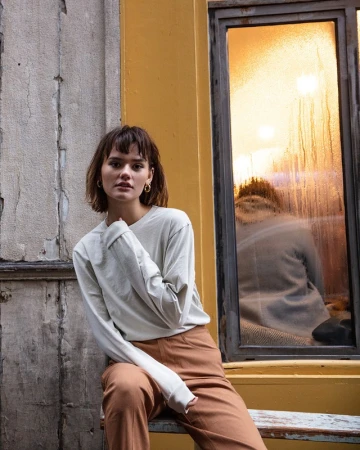 Young adult female with bangs and long-sleeved shirt posing outdoors against concrete wall and yellow-framed window reflecting sunset.
