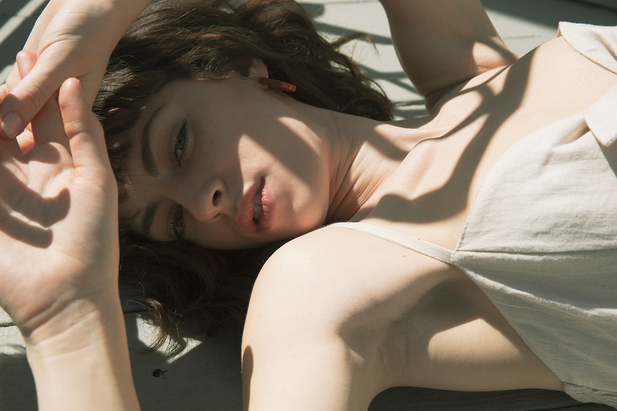 Young woman lying in sunlight wearing a light-colored top, with her arms raised to the side and shadow pattern across her face.