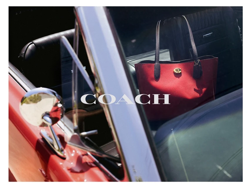 Red leather Coach purse sits on the interior of a classic car, seen through the windshield with the Coach logo on the glass.
