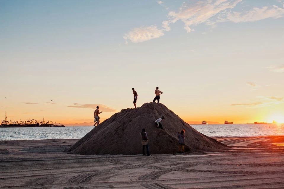 Several young people climb a sand dune at sunset on a beach with the ocean, distant ships, and colorful sky creating a scenic vista.