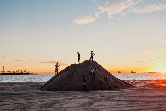 Several young people climb a sand dune at sunset on a beach with the ocean, distant ships, and colorful sky creating a scenic vista.
