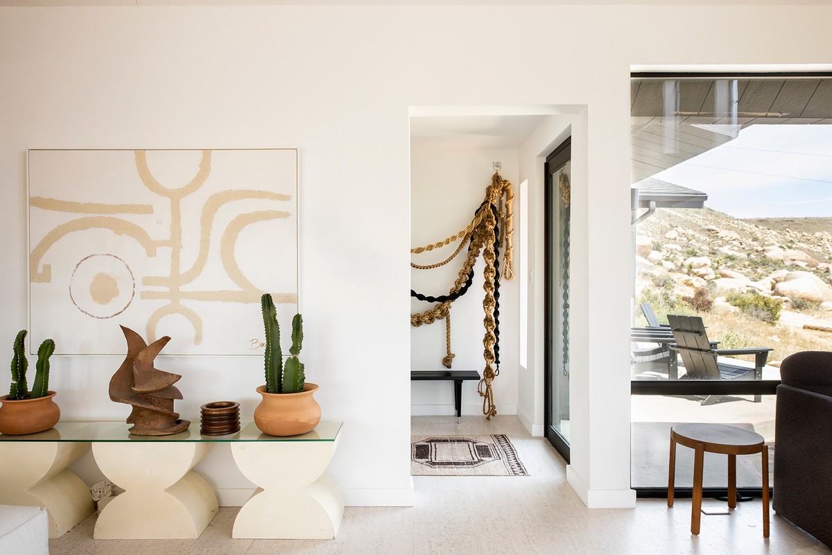 Living room interior featuring abstract art, potted cacti, and a view of a desert landscape through a large window, creating a bright, modern space.